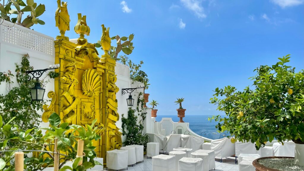 Amalfi Coast terrace with lemon trees, white seating, decorative artwork, and a Mediterranean Sea view.