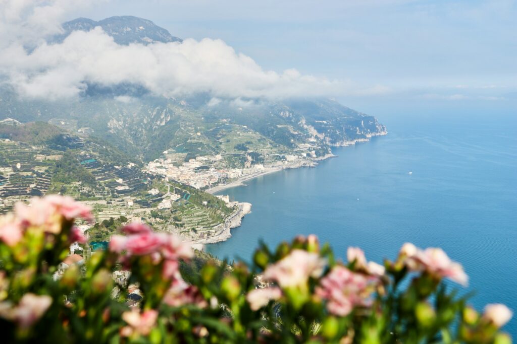 Panoramic Amalfi Coast view with mountains, coastal towns, blue sea, and pink flowers.